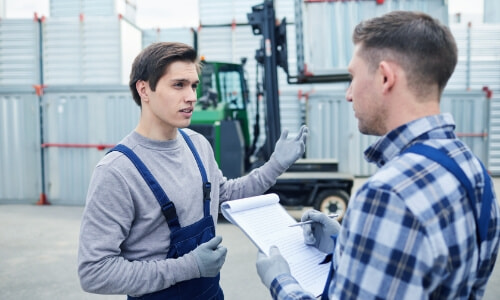 a guy talking to a men with a notepad with a shipping container forklift in the background