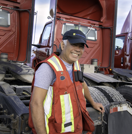 male wearing an orange safety vest and Tag Logistics hat at the back of 3 red semi-trucks